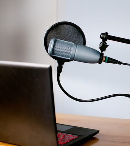 Horizontal image of audio equipment and a laptop sitting on a desk in a small sound room. The studio microphone is on a boom and has a pop filter in place. The laptop is open and ready to use.