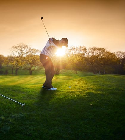 a lone golfer chips onto the green , the low sun is coming from behind him and flaring to camera.