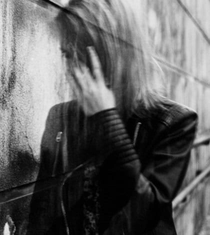 Young woman suffering from a severe disorientation, confusion, or sadness outdoors, in front of a wall. Converted to black and white, grain added, blurry, slightly out of focus.