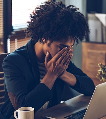 Young African man looking exhausted and covering his face with hands while sitting at his working place