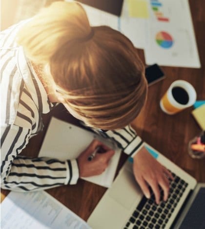 Hardworking successful female entrepreneur working at her desk in her home office writing notes as she researches new markets on the computer, overhead view