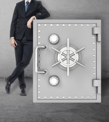 secret box safe guarding 624991814 420 A businessman in a confident pose standing next to the safe on the background of the grey wall. Business and finance. Success and wealth. Protection of funds.