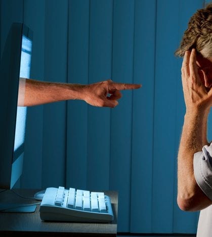 Cyber internet computer bullying and social media stalking Severely distraught young man sitting in front of a computer with a judgmental hand pointing at him from within the computer monitor which shows the man being either computer bullying bullied or Facebook social media stalking stalked.