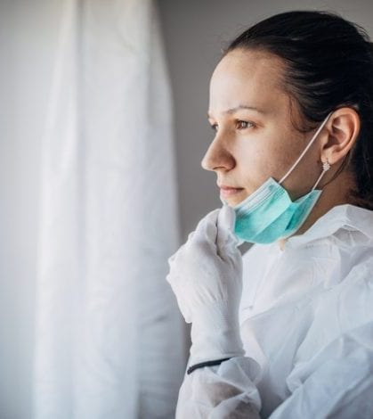 Worried female doctor looking through the hospital window One woman, worried female doctor in protective suit, looking through the hospital window.