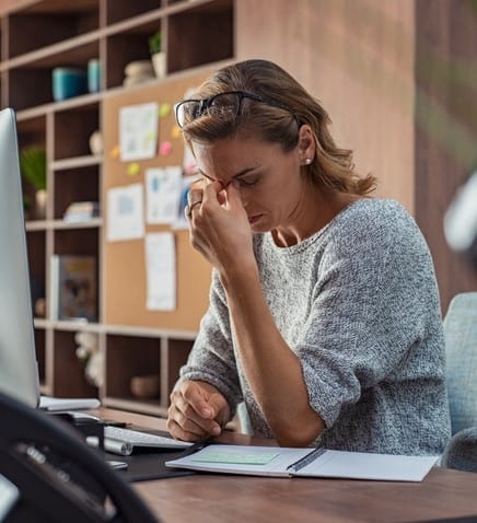 Exhausted businesswoman having a headache in modern office. Mature creative woman working at office desk with spectacles on head feeling tired. Stressed casual business woman feeling eye pain while overworking on desktop computer.