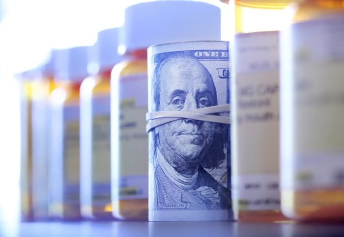 Roll Of One Hundred Dollar Bills In Row Of Prescription Medication Bottles A roll of one hundred dollar bills sits among a row of prescription medication bottles. Photographed with a very shallow depth of field with the focus on the roll of money.