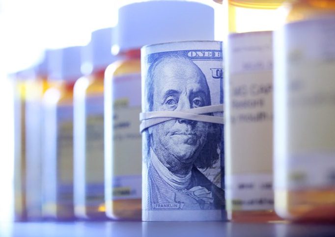 A roll of one hundred dollar bills sits among a row of prescription medication bottles. Photographed with a very shallow depth of field with the focus on the roll of money.