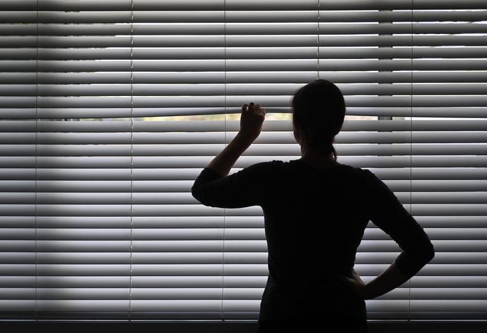 Suspicious woman looking outside through a venetian curtain blinds Suspicious adult woman looking outside home window from behind Venetian curtains. Concept photo of person that have Pistanthrophobia and Agoraphobia, a person who don't trust fear of leaving the house.