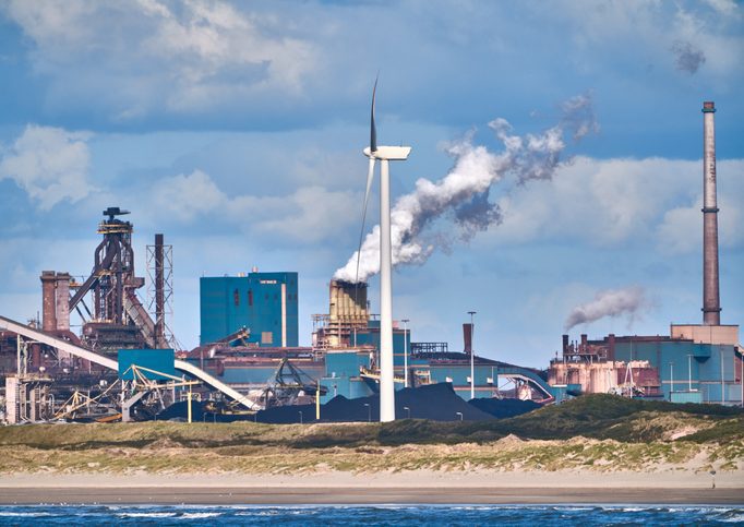 Power generation, wind tower in the foreground, fossil fuel plant with smoke rising in the back.