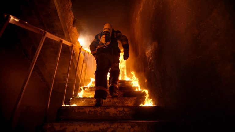 Strong and brave Firefighter Going Up The Stairs in Burning Building. Stairs Burn With Open Flames. Firefighter walking up stairs in a burning building.