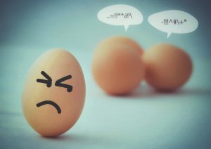 Egg in foreground with frowning face drawn on it, while two eggs in the background, with word balloons, appearing to be gossiping.