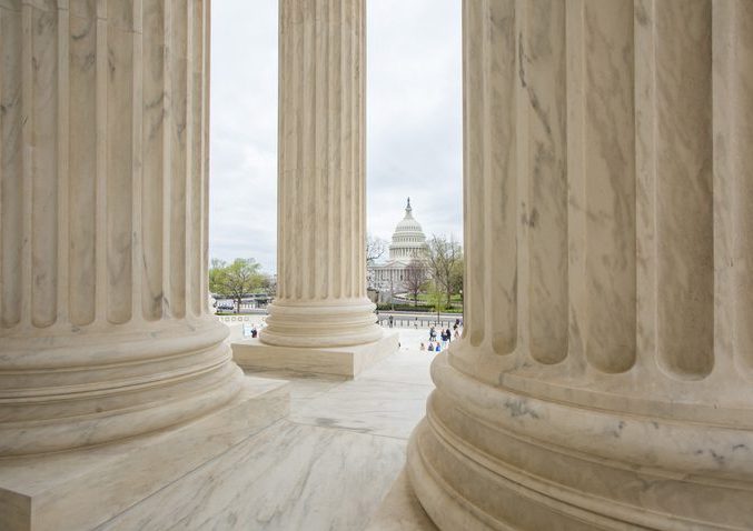 The Capitol Building in DC at a distance, viewed from behind pillars of the Supreme Court.