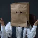 Man wearing suit with bag over his head, shrugging as if he is clueless.