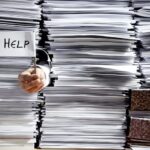 Close-up of stacks of paper, with hand sticking out holding a little sign that says "Help."