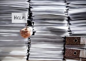 Close-up of stacks of paper, with hand sticking out holding a little sign that says "Help."