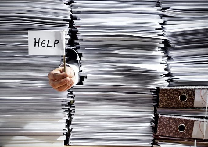 Businessman's Hand Holding Help Flag And Sticking Out Of Paper Close-up of stacks of paper, with hand sticking out holding a little sign that says "Help."