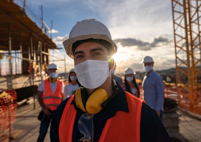 Woman construction worker in foreground, wearing a Covid mask, with construction site and other workers in the background.