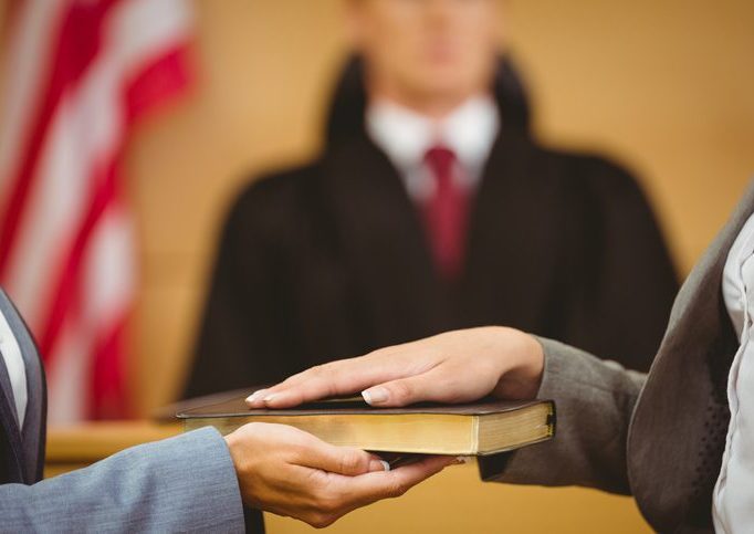 Close-up, hands, someone talking an oath on a Bible or book, with a judge and flag in the background.