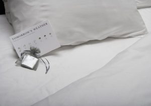 Close-up of a pillow with a chocolate and weather report card next to it, as if in a hotel room.