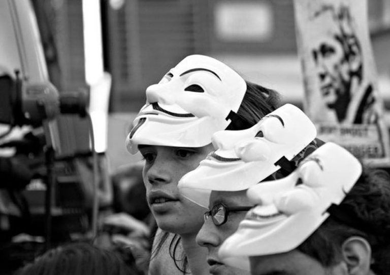 subterfuge-fake-deception-Assange-demo-466824560-e1660917648293 Demonstrators with masks at a demonstration in support of Assange, his photo faintly visible on a sign in the background.