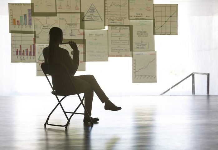 Businesswoman studying charts and graphs on wall in office Business person sitting on a folding chair looking at a wall hung with charts and graphs.