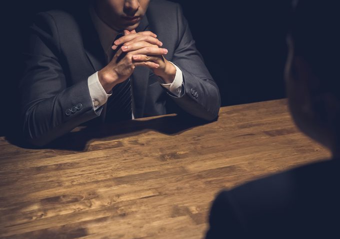 Detective interviewing suspect in dark room Dark image of a man in a suit sitting with his hands crossed at a table, before a not quite visible person across from him who seemingly is being called to account.