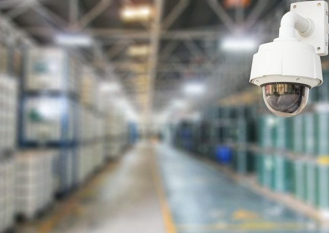 Close-up of a 360 degree installed camera with a long warehouse aisle out of focus in the background.
