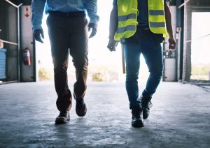 Two men walking through a warehouse, one man in a dress shirt the other with a worker's high-visibility vest.