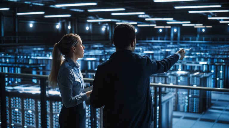 Data Center Male It Specialist and Female e-Business Fintech Startup Manager Talk, Use Laptop. Cloud Computing Server Farm with Two Information Technology Professionals working. Two young techies looking out over what appears to be a bank of servers.