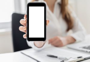 Person behind a desk holding up a smartphone, with screen in the direction of the viewer.