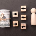 An symbolic assembly of objects: On the left a roll of bills; in the middle tiles with arrows on them, some pointing left, some right; on the right a wooden piece, a bit like a chess piece. Symbolically, that figure could be "liable."