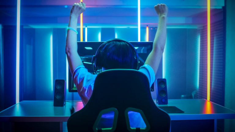 Viewed from the back, a young boy, wearing earphones, engrossed in a computer screen, arms raised in triumph.