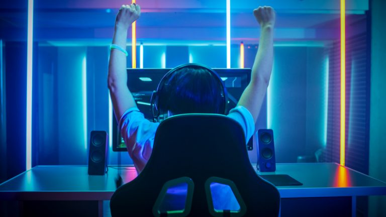 Professional Gamer Playing and Winning in First-Person Shooter Online Video Game on His Personal Computer. Footage Fade out into Bokeh. Room Lit by Neon Lights in Retro Arcade Style. Cyber Sport Championship. Viewed from the back, a young boy, wearing earphones, engrossed in a computer screen, arms raised in triumph.