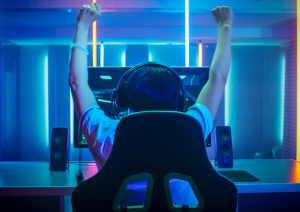 Viewed from the back, a young boy, wearing earphones, engrossed in a computer screen, arms raised in triumph.