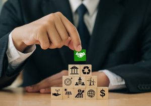 Front view, man wearing suit and tie at a table, making a pile of block, each of which has an illustration. The top block he is setting is green. and has illustration of a hand shake.