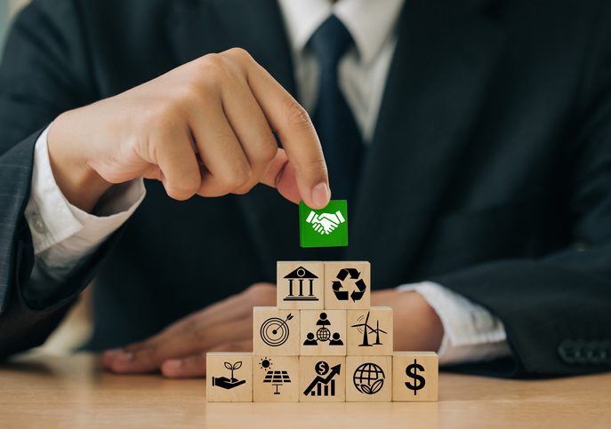 Businessmen holding green Wooden blocks with handshake icons standing with other ESG icons idea for Business cooperation for environmental, social, and governance. Sustainable corporation development. Front view, man wearing suit and tie at a table, making a pile of block, each of which has an illustration. The top block he is setting is green. and has illustration of a hand shake.
