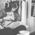 Black & white photo of a nursing mother sitting comfortably in what looks like it could be storage closet.