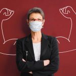 Woman wearing mask facing forward, arms folded. The background is a superimposed sketch of a muscular arms flexing biceps.