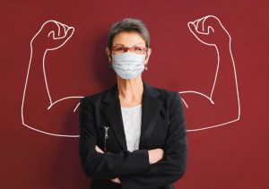 Woman wearing mask facing forward, arms folded. The background is a superimposed sketch of a muscular arms flexing biceps.