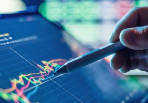 Close up rendering of a hand holding a marker over a graph with a number of colored lines already drawn on it.