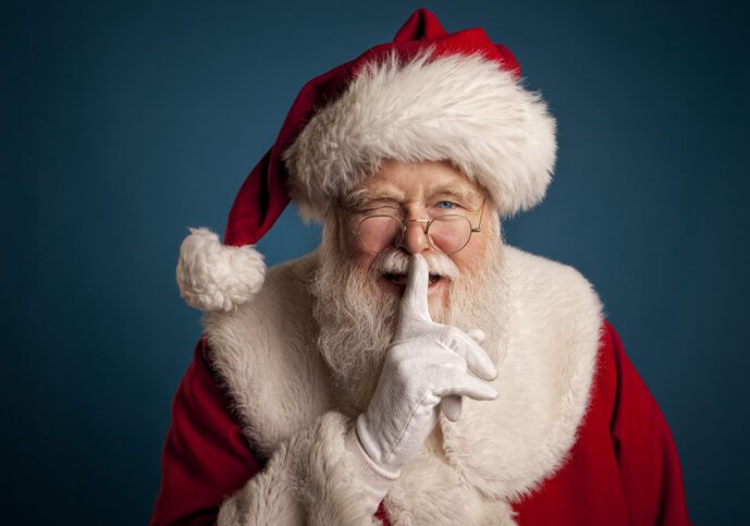 Man in Santa Claus costume looking at viewer, with his finger over his mouth as of to say "shhh."