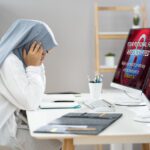 Woman with possibly a head covering, presumably a health care staff person, sitting and looking distraught before a computer screen with a not quite legible but alarming looking message on the screen.