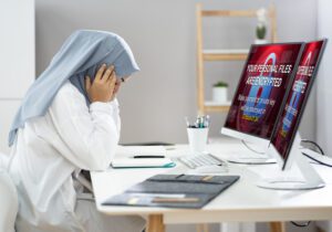 Woman with possibly a head covering, presumably a health care staff person, sitting and looking distraught before a computer screen with a not quite legible but alarming looking message on the screen.