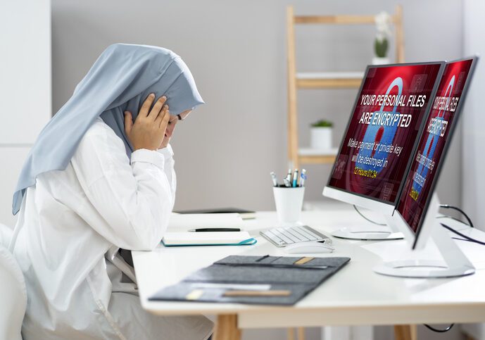 Ransomware Cyber Attack Woman with possibly a head covering, presumably a health care staff person, sitting and looking distraught before a computer screen with a not quite legible but alarming looking message on the screen.