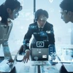 Three business people gathered around a laptop on a table, with vague technology-suggesting icons superimposed over the blue-tinged photo. One of the persons appears to be explaining something to the other two.