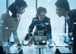 Three business people gathered around a laptop on a table, with vague technology-suggesting icons superimposed over the blue-tinged photo. One of the persons appears to be explaining something to the other two.