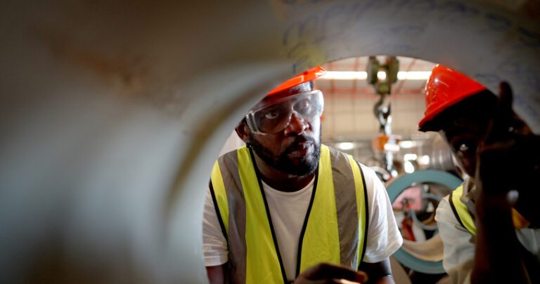 Close up. Professional African American industrial engineer or worker or foreman in metal work, wear personal protective equipment, monitor roll steel the raw material store in production line at metal part manufacturing factory. Two inspectors, hearing orange hard hats and yellow vests, are seen looking into a large pipe, viewed from inside the pipe.