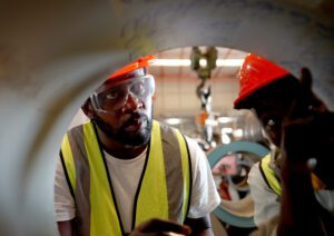 Two inspectors, hearing orange hard hats and yellow vests, are seen looking into a large pipe, viewed from inside the pipe.