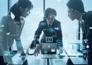 Three business people gathered around a laptop on a table, with vague technology-suggesting icons superimposed over the blue-tinged photo. One of the persons appears to be explaining something to the other two.
