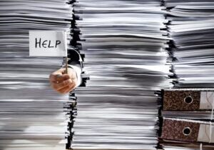 Close-up of stacks of paper, with hand sticking out holding a little sign that says "Help."
